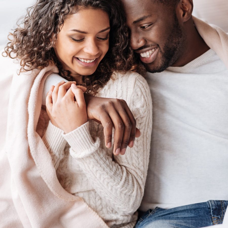 Happy to be with you. Smiling young cheerful African American couple sitting in the cafe and being covered with a blanket while expressing care and hugging